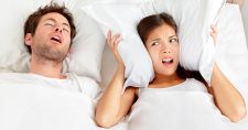 Snoring? Trouble Sleeping? Try Exercise. Picture of a caucasian male sleeping in bed and a caucasian female holding her pillow around her ears.